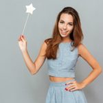 Cheerful beautiful young woman holding magic wand over grey background